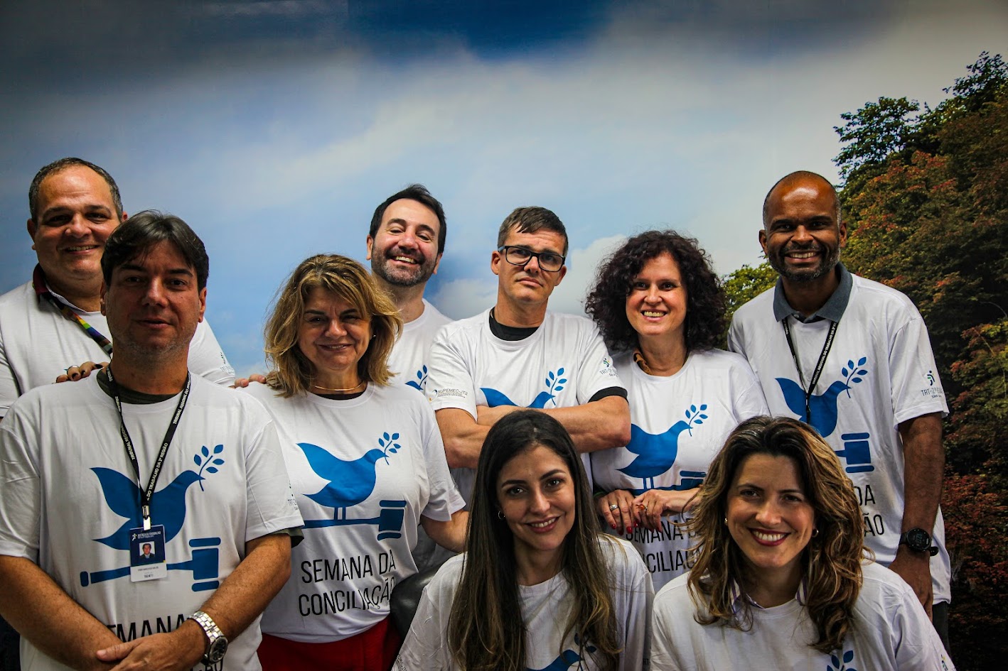 A imagem apresenta um grupo de nove pessoas, com a maioria usando camisetas brancas estampadas com um desenho de uma pomba azul e martelos de juiz, e algumas com o texto