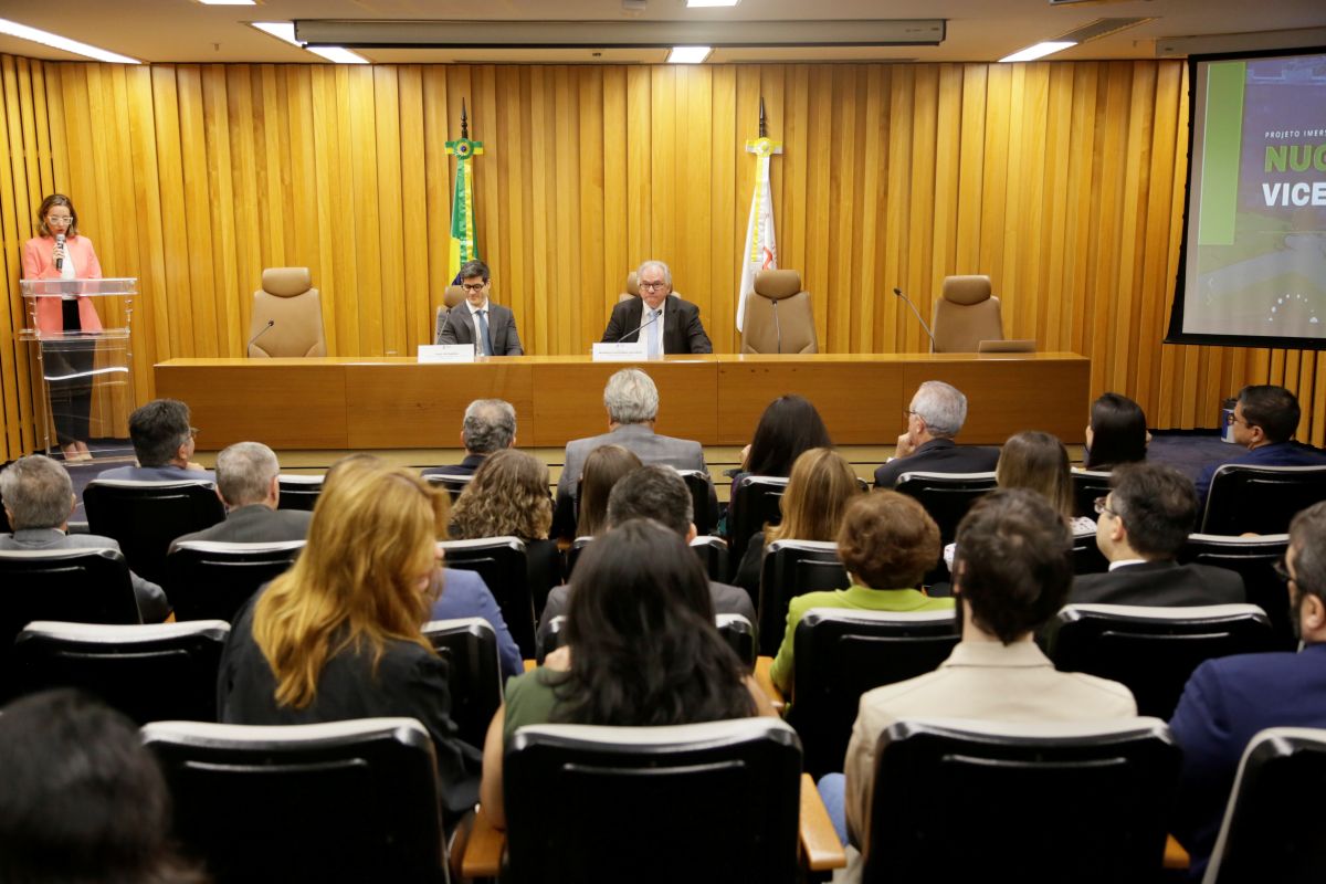 Mesa de abertura do evento. Auditório pequeno, porém, cheio. Cadeiras lotadas e duas pessoas na mesa de abertura.