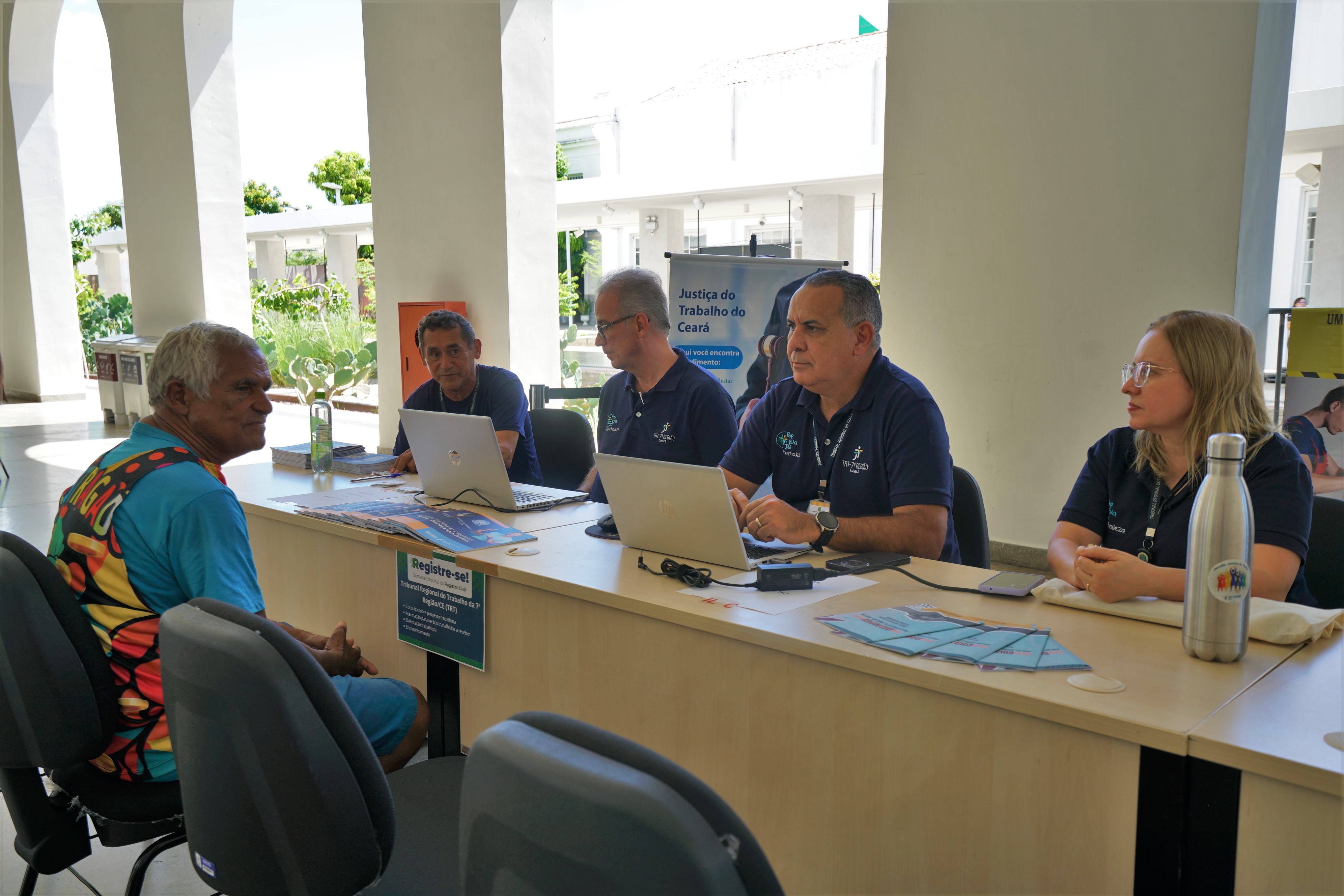 Equipe da Justiça do Trabalho do Ceará atende um homem em um balcão de serviço, com computadores e materiais informativos, em ambiente claro e aberto.