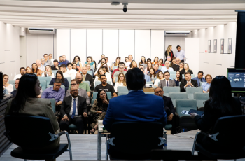 A imagem mostra um auditório cheio, com especialistas e estudantes assistindo a um painel. Três pessoas estão sentadas de costas, conduzindo o evento.