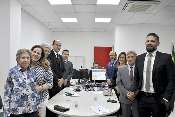 A imagem mostra um grupo de pessoas reunidas em uma sala de reuniões. Elas estão de pé ao redor de uma mesa branca com computadores, papéis e copos descartáveis.