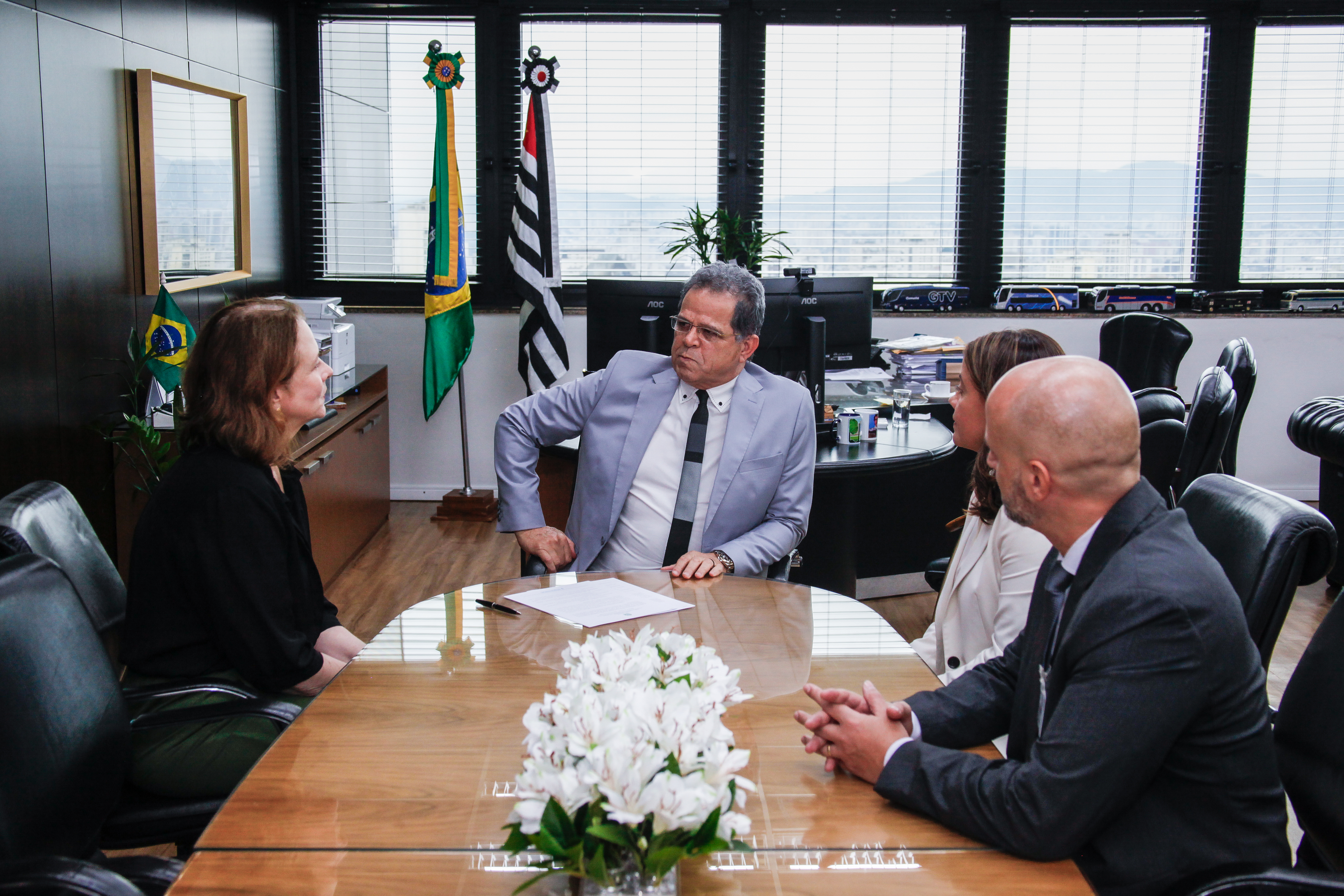 Quatro pessoas estão sentadas em uma mesa de reunião, conversando. Ao fundo, há as bandeiras do Brasil e do estado de São Paulo, além de janelas grandes que mostram a vista externa.