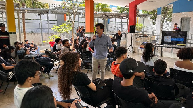 Fotografia com a presença de um homem com microfone na mão e algumas pessoas ao lado sentadas.