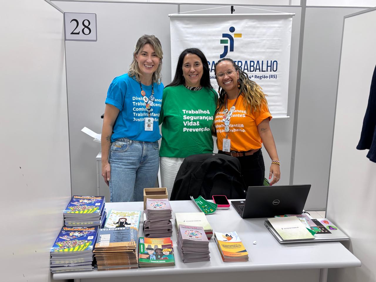 Fotografia com a presença de 3 mulheres e a frente alguns conjuntos de revistas, notebook e celular. 