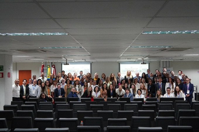 Fotografia com diversas pessoas ao fundo de uma sala com cadeiras pretas posicionadas lado a lado.