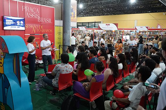 Imagem de uma feira de livros, com pessoas sentadas assistindo a uma apresentação na “Praça Assis Brasil”. Dois palestrantes estão em pé na frente, falando ao público. Ao fundo, há estandes de livros e várias pessoas circulando.