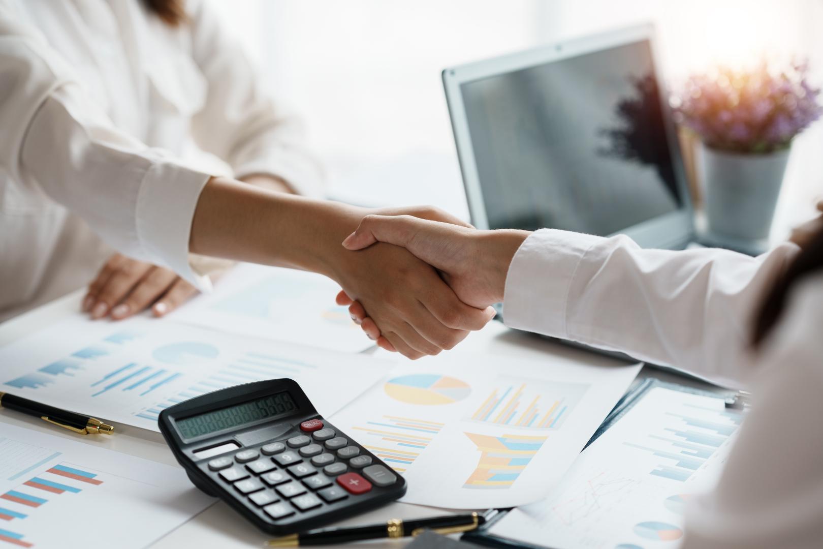 Fotografia com aperto de mãos entre duas pessoas. Ao lado, papeis, computador, canetas e calculadora em cima da bancada. 