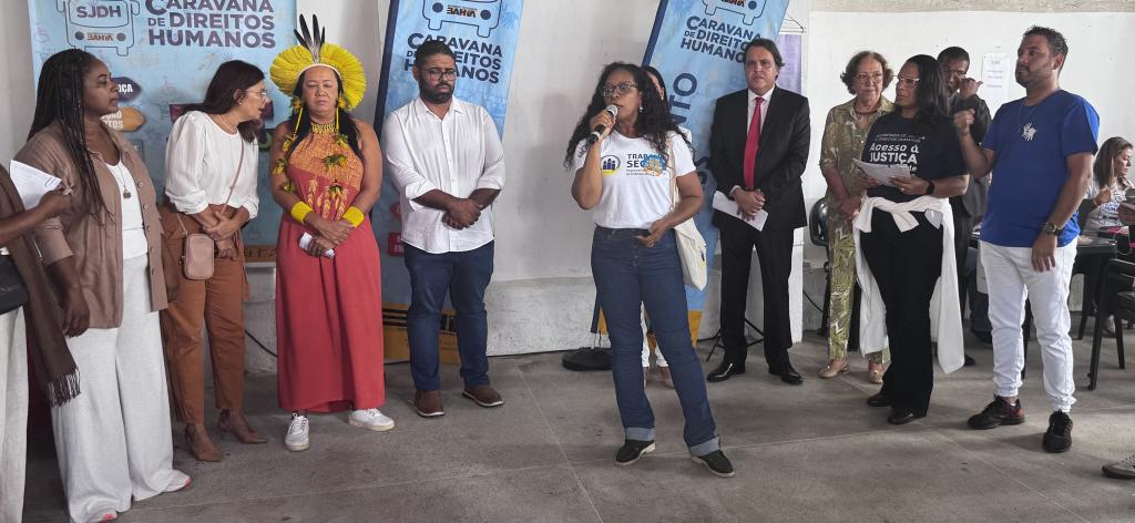 A imagem mostra um grupo de pessoas reunidas em um evento da Caravana de Direitos Humanos. Uma mulher fala ao microfone, enquanto os demais escutam. Ao fundo, há banners do evento e todos estão em um ambiente coberto.