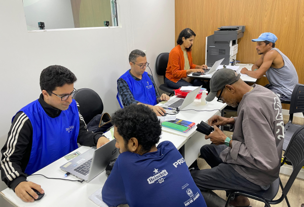 A imagem mostra uma sala com atendentes usando coletes azuis auxiliando pessoas em computadores, possivelmente em um mutirão de serviços sociais ou emissão de documentos.