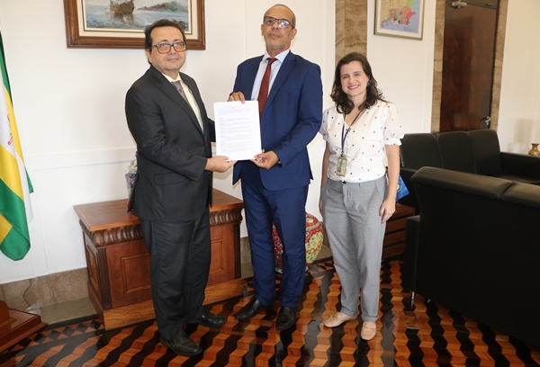 Duas pessoas em traje formal seguram um documento, ao lado de uma mulher sorridente.