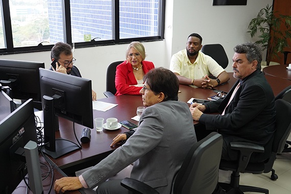 Fotografia de uma mesa com algumas pessoas sentadas com o corpo virado para um computador. 