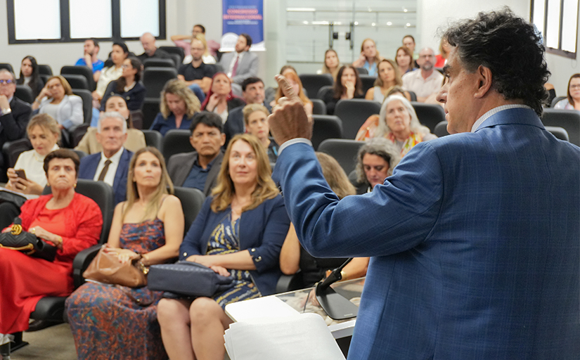 Foto de um homem de costas, de terno, dando uma palestra. Ao fundo o público assistindo.