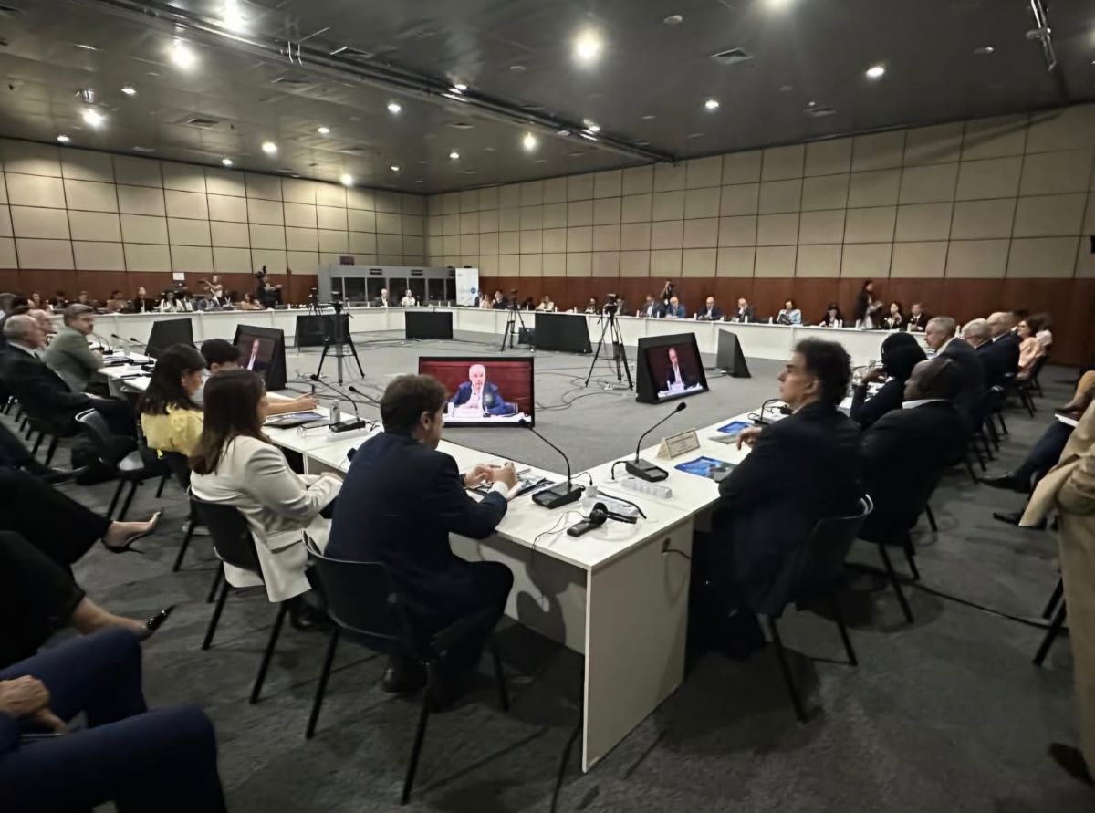 Foto do evento. Uma grande mesa quadrada com diversas pessoas sentadas.