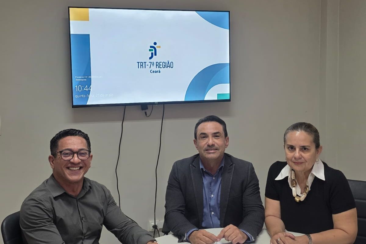 Três pessoas estão sentadas lado a lado em uma sala de reuniões, posicionadas em frente a uma TV fixada na parede que exibe o logotipo do "TRT-7ª REGIÃO Ceará" e o relógio marcando 10:44. À esquerda, um homem sorridente de óculos veste uma camisa social cinza-escura. No centro, outro homem usa camisa azul sob um paletó escuro, e à direita, uma mulher veste uma blusa preta com gola branca e um colar de pedras claras.