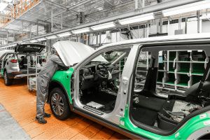 A imagem mostra um operário uniformizado trabalhando na parte frontal de um veículo prata em uma linha de montagem automotiva moderna e iluminada.