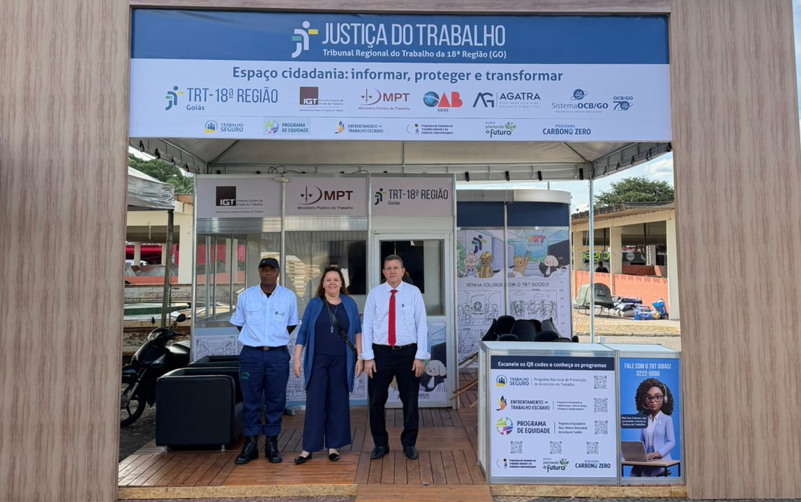 Fotografia da fachada do espaço “Justiça do Trabalho”, montado em estrutura temporária de atendimento ao público. Na entrada, três pessoas posam para a foto: um homem de camisa clara e boné, uma mulher de roupa azul e um homem de camisa branca com gravata vermelha. Acima da estrutura, há uma placa azul com a inscrição “Justiça do Trabalho – Espaço cidadania: informar, proteger e transformar”, além de logomarcas de instituições parceiras. Cartazes informativos estão expostos nas laterais da entrada.