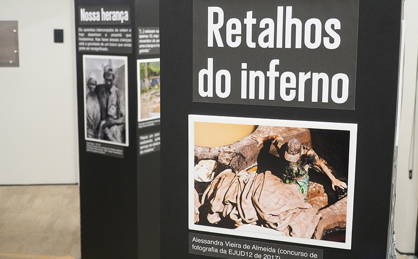 Totem de uma exposição mostrando a foto de um trabalhador no meio de muitos tecidos. O texto acima da foto é 