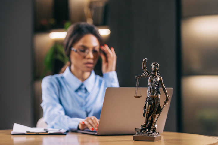 Mulher ajeita o óculos diante de computador e estátua da Justiça, com documentos sobre a mesa