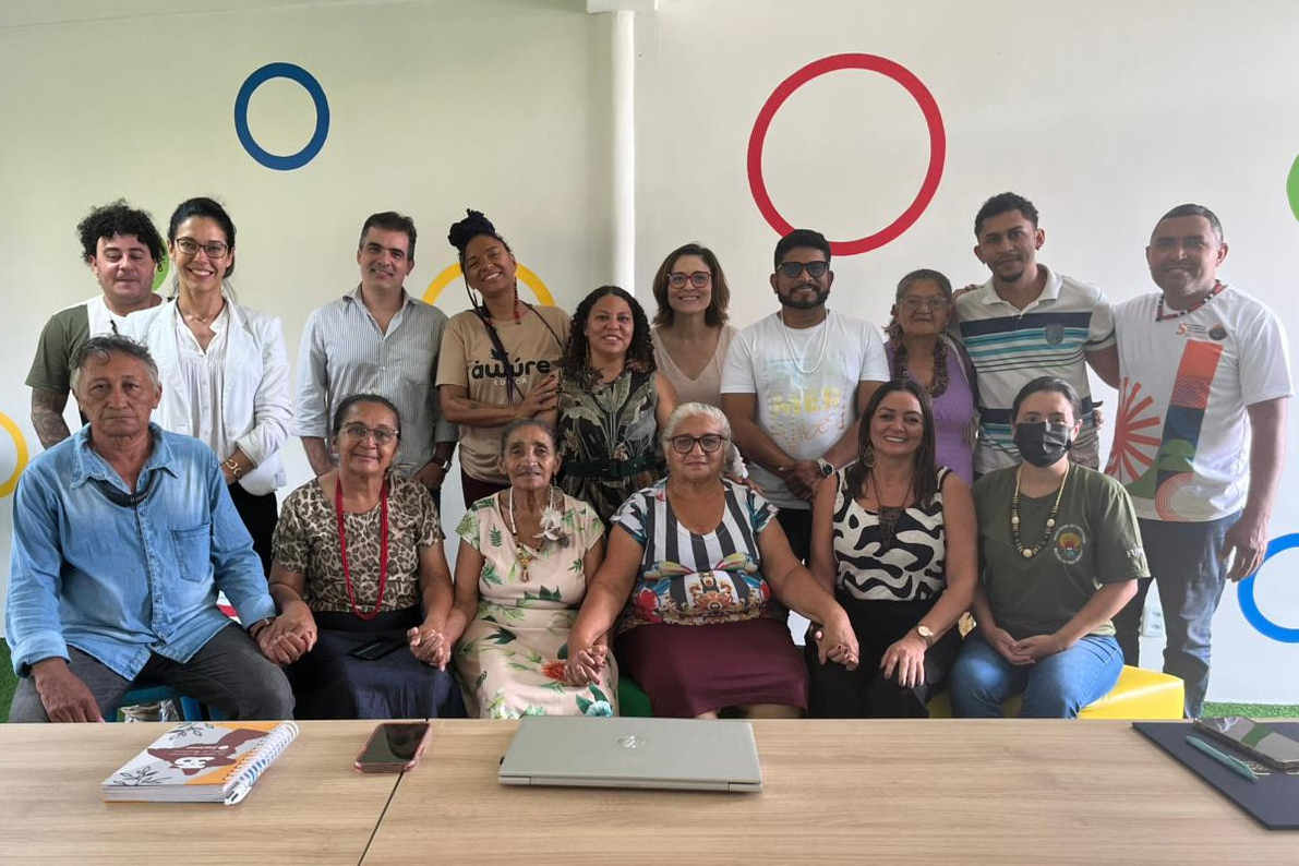 Grupo de pessoas reunidas, algumas sentadas e outras em pé, sorrindo para a foto em um ambiente interno com decoração colorida na parede.