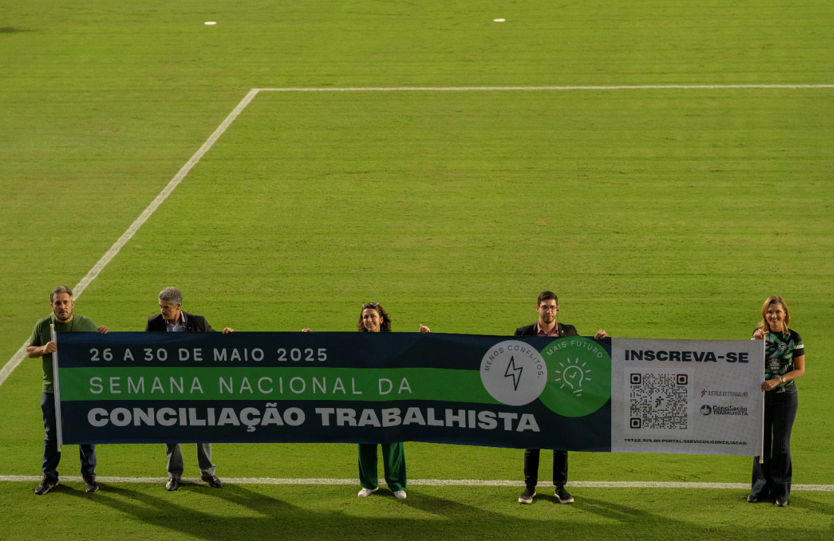 Cinco pessoas seguram uma faixa horizontal extensa com a divulgação da Semana da Conciliação Trabalhista, atrás dela, o gramado verde de um campo de futebol.