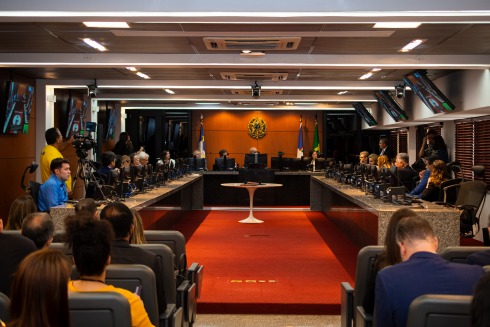 A imagem mostra uma sessão formal em um tribunal, com várias pessoas sentadas em cadeiras, juízes ao fundo e bandeiras do Brasil e da Bahia.