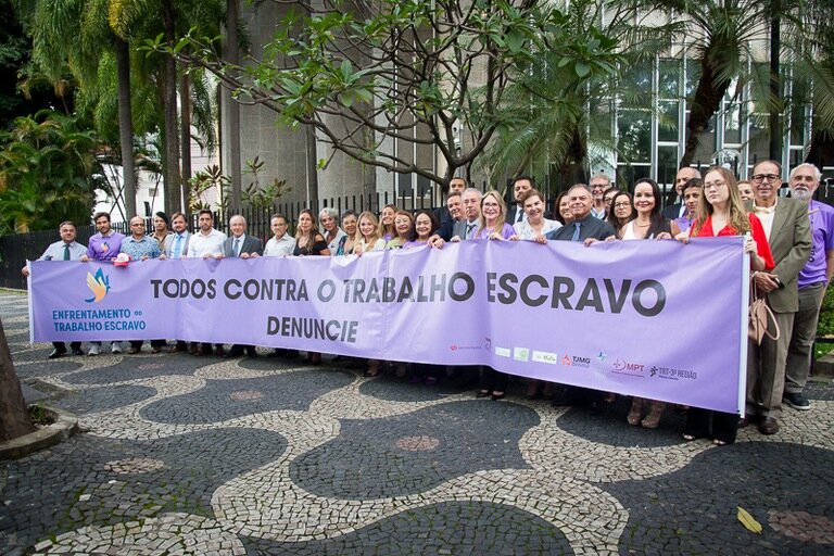 Fotografia com um conjunto de pessoas mobilizadas com um cartaz roxo em mãos e a escrita ''Todos Contra o Trabalho Escravo Denuncie''.
