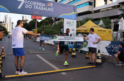 Fotografia de um homem ultrapassando a faixa referente a 7ª Corrida do Trabalho. 