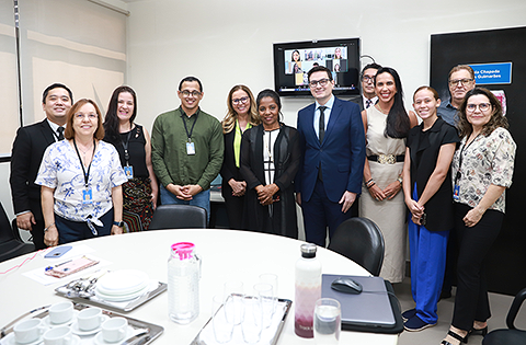 Fotografia com 7 mulheres e 5 homens em foco, todos com roupa social, e em frente à uma mesa com garrafas, copos e um computador.