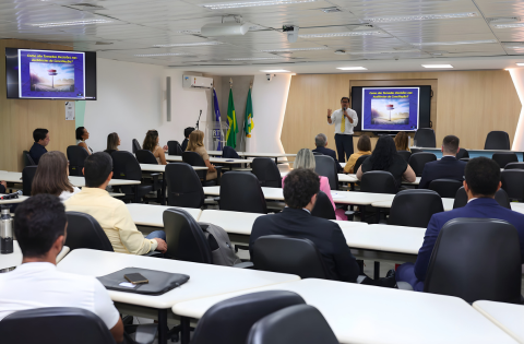 Fotografia de uma palestra com algumas pessoas sentadas assistindo e um palestrante em pé ao lado de um banner.