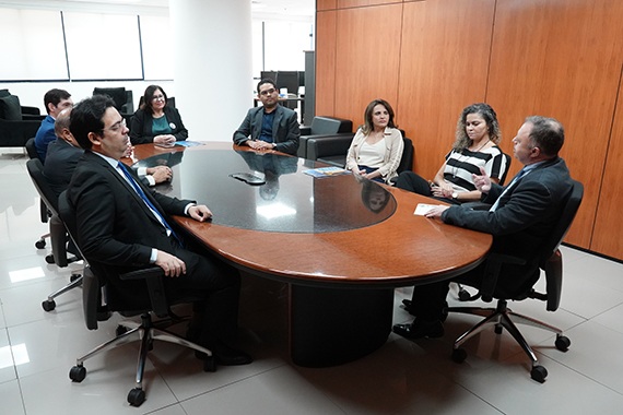 A imagem mostra um grupo de pessoas em reunião ao redor de uma mesa oval de madeira.