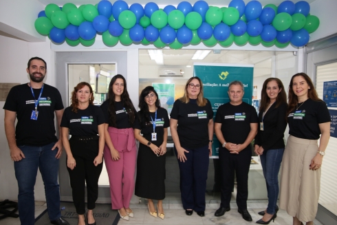 A imagem mostra um grupo de nove pessoas posando para foto em frente a uma sala decorada com balões verdes e azuis. Todos usam camisetas pretas com a mesma estampa, indicando participação em um evento ou campanha institucional.