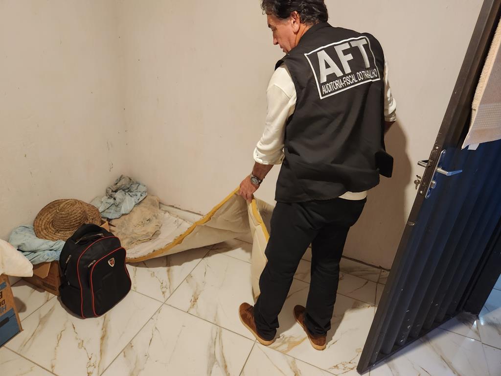 Auditor-fiscal do trabalho inspeciona um cômodo precário, com colchão improvisado no chão e poucos pertences pessoais.
