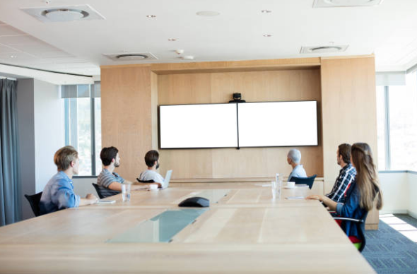 Grupo de pessoas reunidas em uma mesa em uma sala com duas telas para videoconferências 