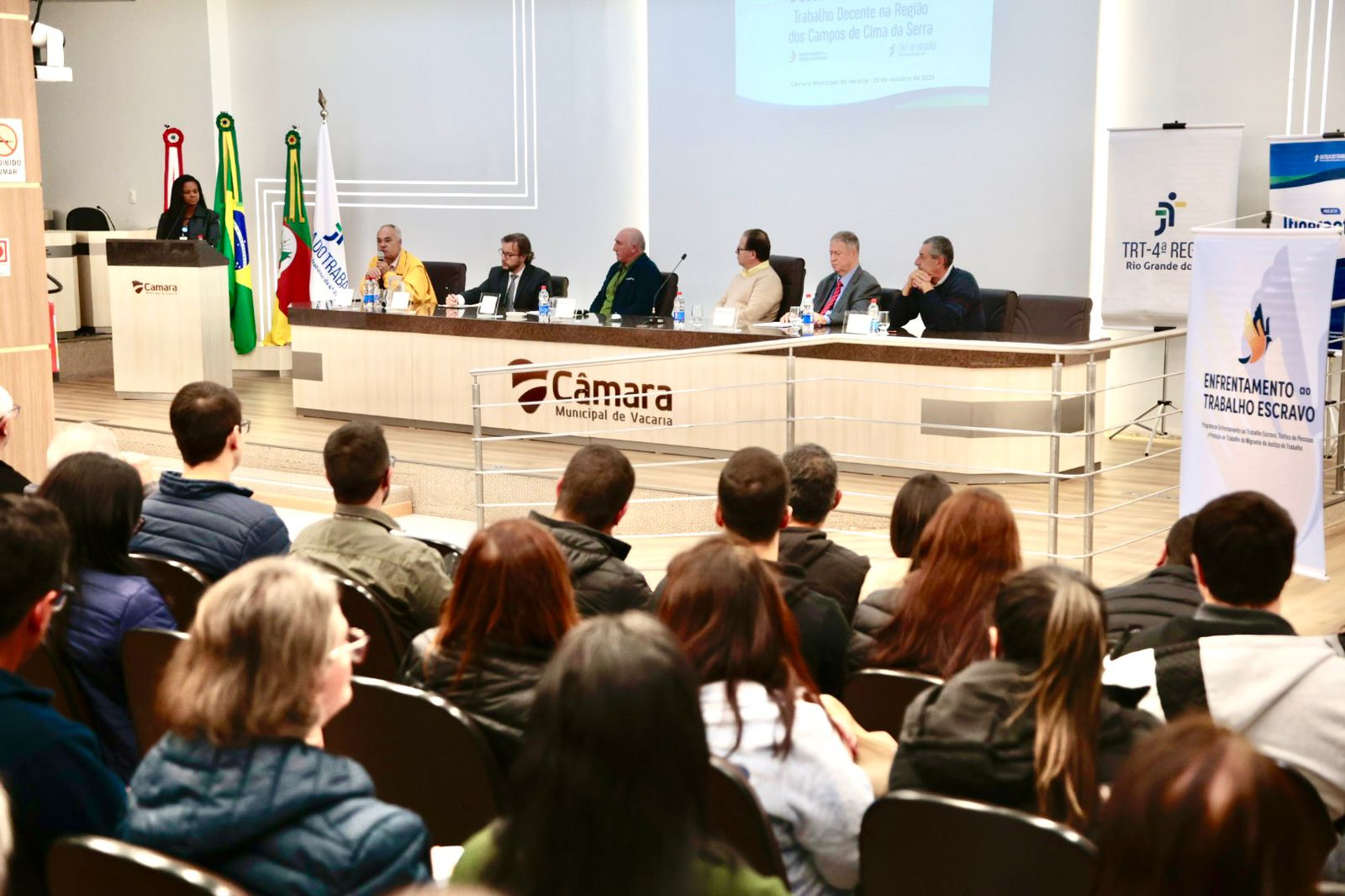 Fotografia de uma mesa diretora e de um público sentado; ao lado alguns cartazes e acima deles um slide projetado.