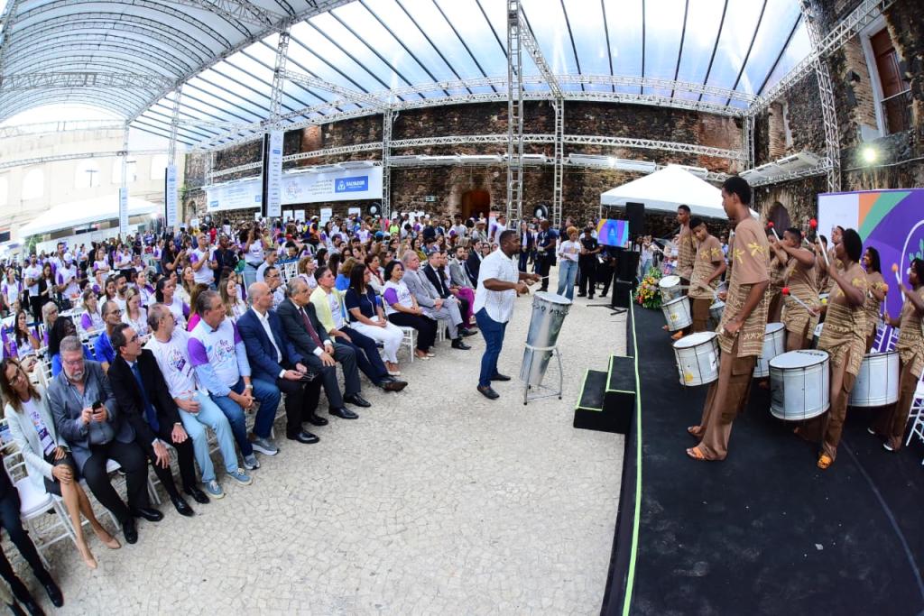 Fotografia com a presença de diversas pessoas na plateia e um grupo se apresentando com alguns instrumentos e um maestro para reger o grupo.