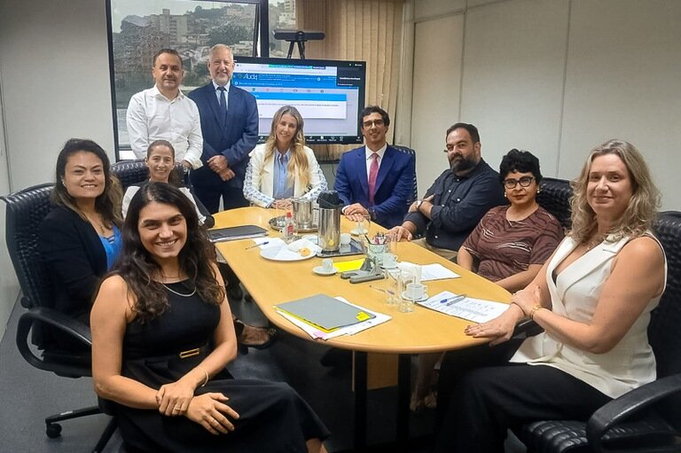Fotografia com 10 pessoas, sendo 6 mulheres e 4 homens, ao redor de uma mesa e em frente à tela de um computador. 