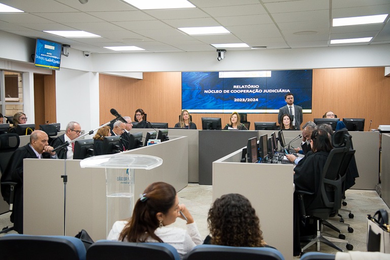 Fotografia com a presença de diversas pessoas e o banner ''Relatório Núcleo de Cooperação Judiciária 2023/2024'' ao fundo.