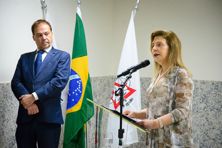 Mulher discursa em um púlpito com microfone, enquanto um homem de terno a observa ao fundo. Atrás deles, estão as bandeiras do Brasil e do TRT da 17ª Região. Cena ocorre em ambiente formal e institucional.