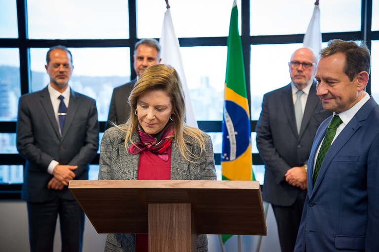 A imagem mostra uma mulher assinando um documento em um púlpito, com quatro homens ao fundo observando. Ao centro, há uma bandeira do Brasil e duas bandeiras brancas. Todos estão vestidos formalmente, em ambiente interno e institucional.