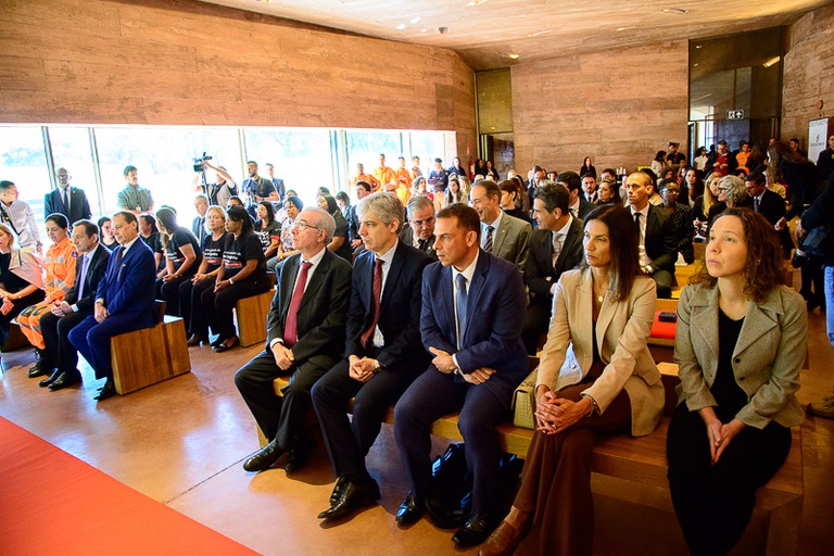 Um grupo de pessoas, vestidas formalmente, está sentado em bancos de madeira dentro de um auditório ou salão, assistindo a um evento.