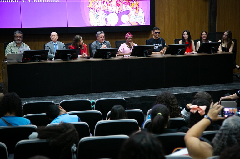 Fotografia com a presença de 9 pessoas em uma bancada diretora, sendo 5 mulheres e 4 homens. 