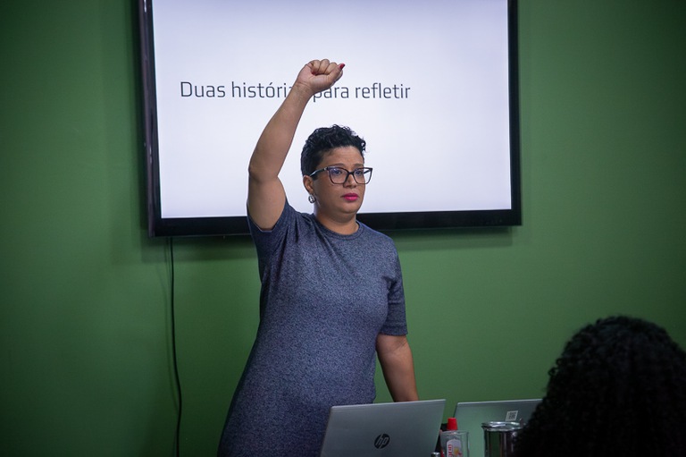Fotografia de uma mulher com um braço levantado e atrás a tela de um computador com a legenda ''Duas histórias para refletir''. 