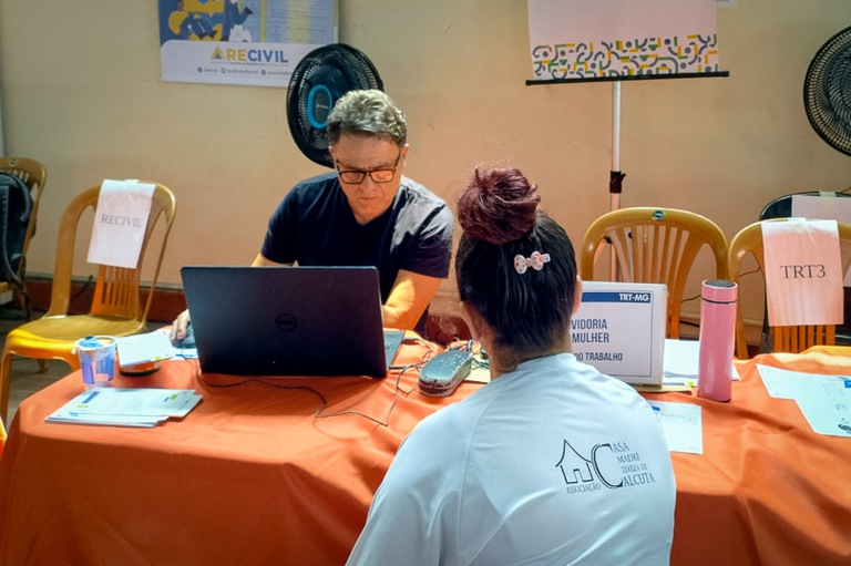 Fotografia com a presença de um homem em frente a um notebook, uma mulher de costas e uma mesa com objetos.