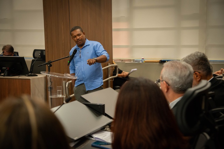 Fotografia com a presença de um homem com blusa azul em frente a microfone, discursando algo para algumas pessoas que estão sentadas. 