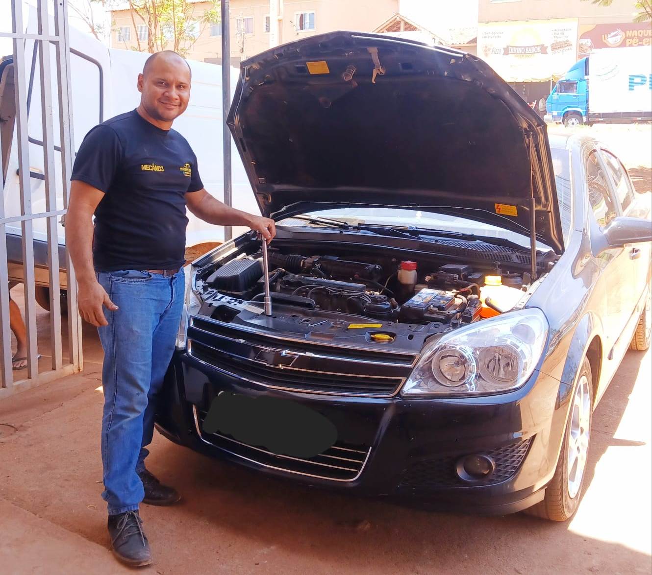 O venezuelano Emílio posando para foto na frente de um carro de capô aberto. Ele é mecânico.