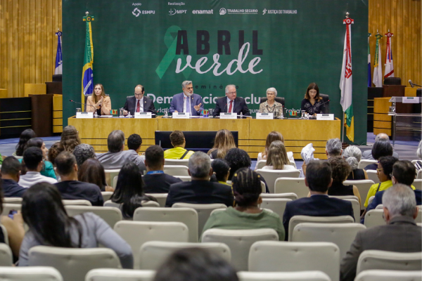 Foto do plenário com pessoas sentadas nas cadeiras e outras na mesa de exposição. Ao fundo, um backdrop escrito Abril Verde.